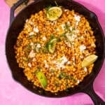 A large skillet pan with golden browned sweet corn topped with crumbled feta, lime wedges and fresh coriander on a pink background.