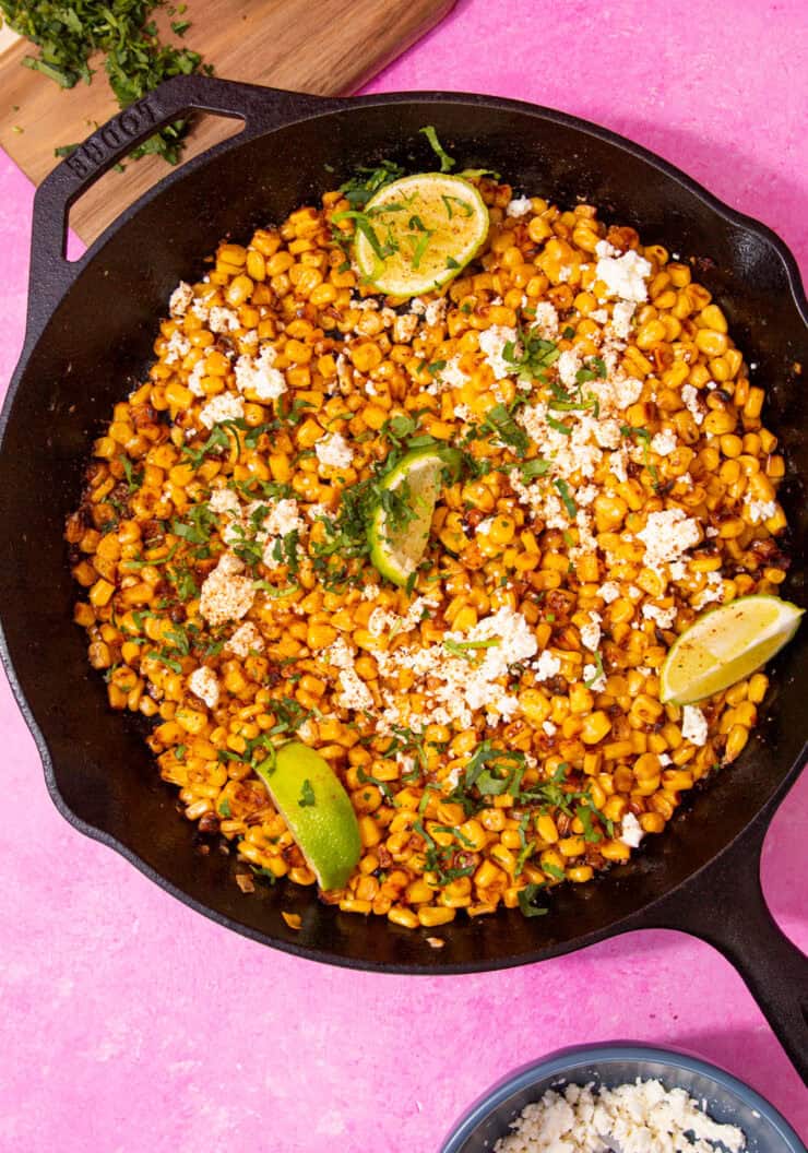 A large skillet pan with golden browned sweet corn topped with crumbled feta, lime wedges and fresh coriander on a pink background.