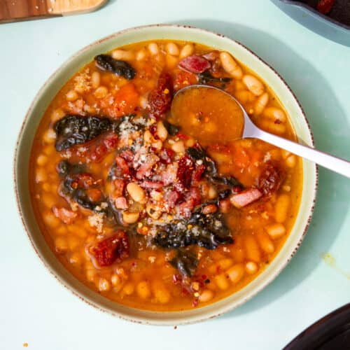 A bowl of white bean soup with cavolo Nero, sundried tomatoes and crispy bacon bits and a spoon.