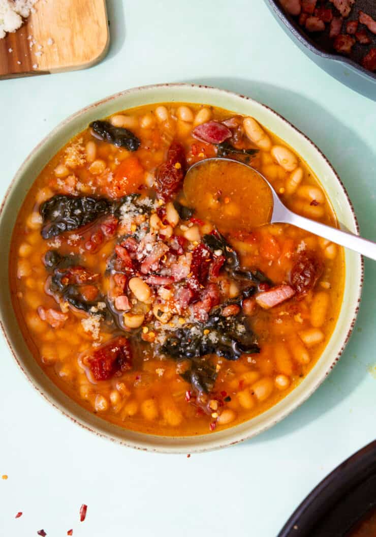 A bowl of white bean soup with cavolo Nero, sundried tomatoes and crispy bacon bits and a spoon.