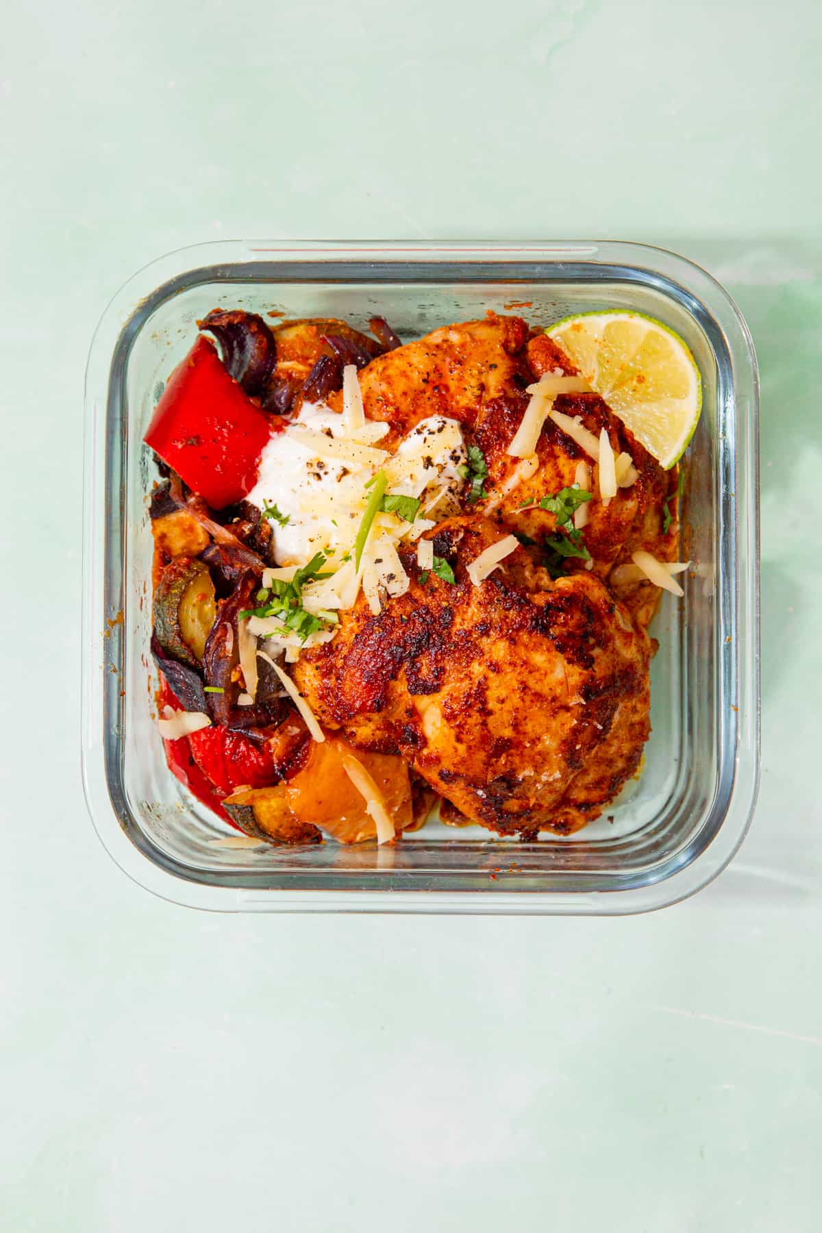 1 meal prep square, glass meal prep container with roasted chicken, vegetables, sour cream, a wedge of lime and grated cheddar.