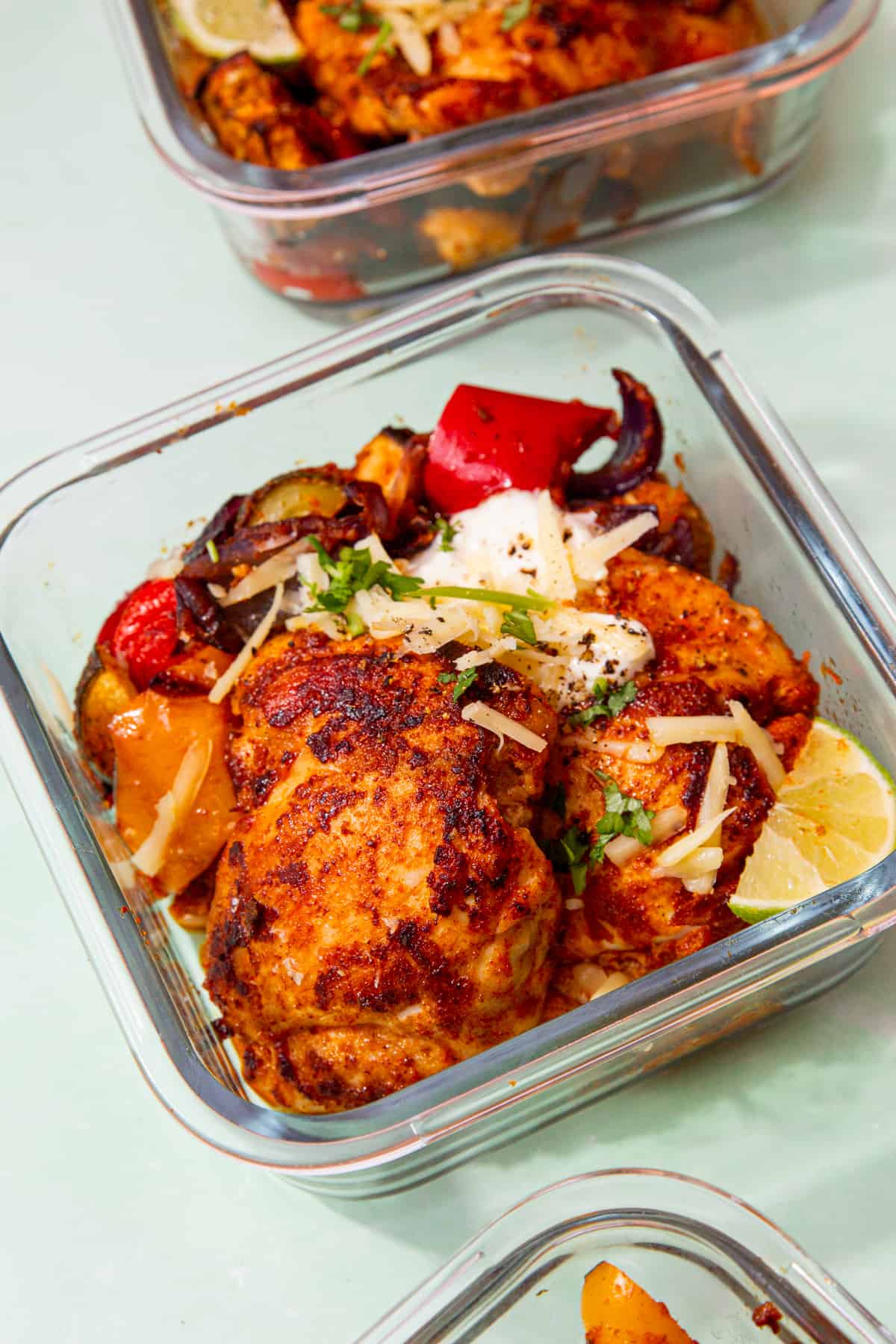 A few square, glass meal prep container with roasted chicken, vegetables, sour cream, a wedge of lime and grated cheddar.