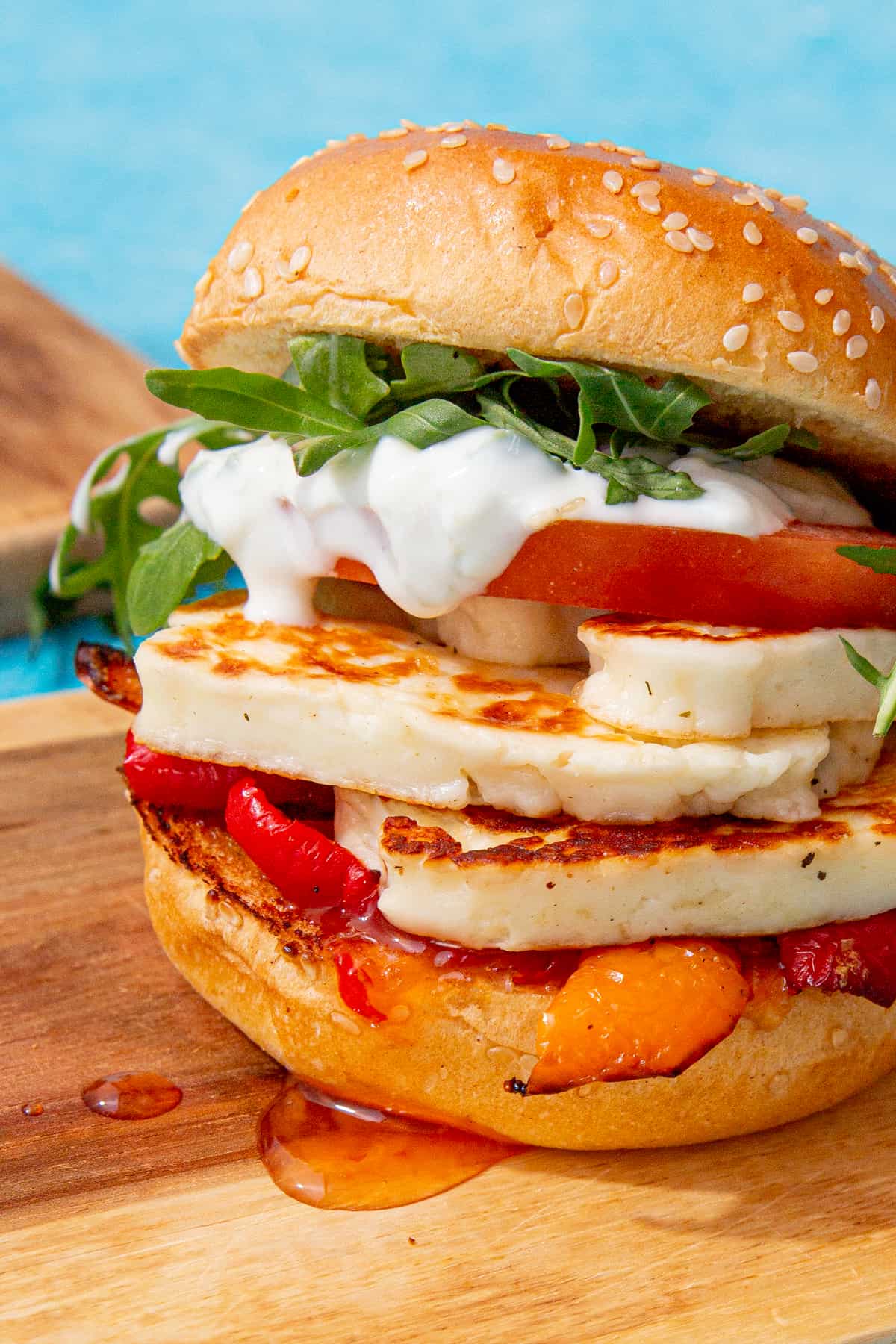 A burger with golden browned halloumi slices, peppers, tomatoes rocket, sweet chilli sauce and tzatziki in a sesame bun.