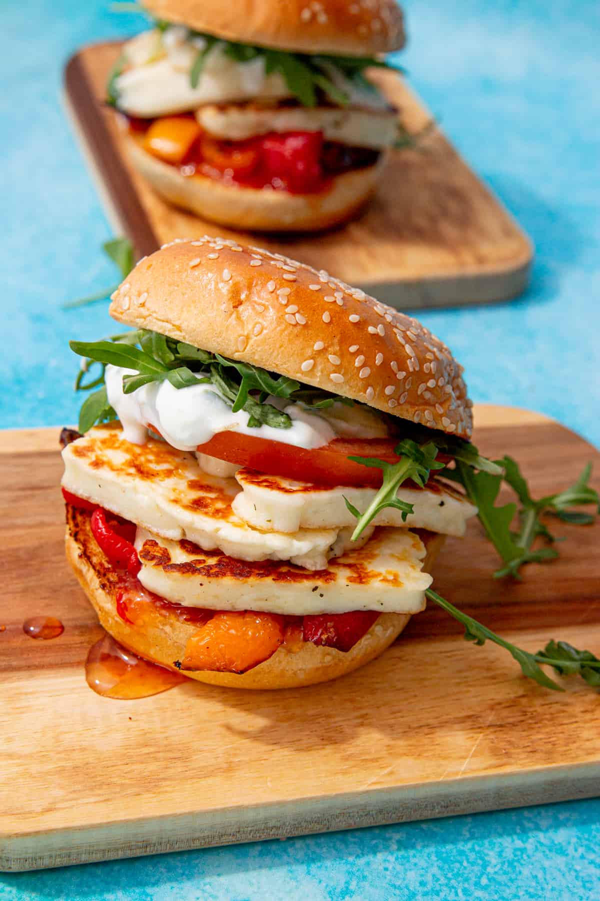 2 halloumi burgers with golden browned halloumi slices, peppers, tomatoes rocket, sweet chilli sauce and tzatziki in a sesame bun on 2 mini wooden chopping boards.
