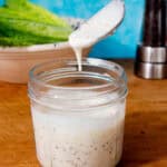 A glass container with dressing and a spoon pouring some dressing in with lettuce in partial view behind.