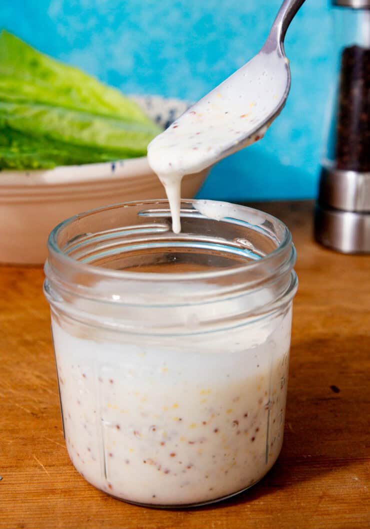 A glass container with dressing and a spoon pouring some dressing in with lettuce in partial view behind.