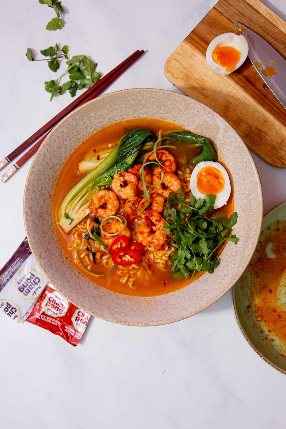 A bowl of prawn ramen with pak choi, coriander, chillies, half an egg in the broth next to some chop sticks and the other half of the egg on a chopping board. 