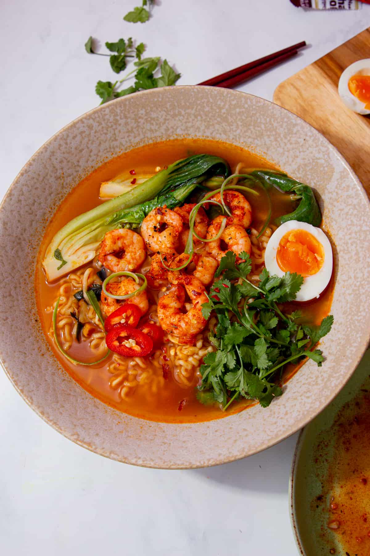 A bowl of prawn ramen with pak choi, coriander, chillies, half an egg in the broth next to some chop sticks and loose coriander and the other half of the egg on a chopping board. 