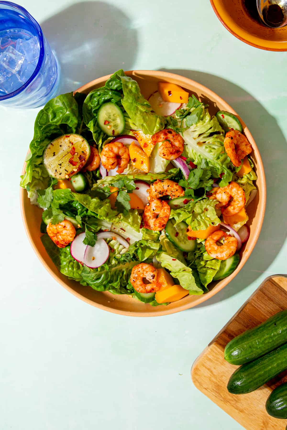 Lots of golden browned king prawns in a bowl of salad with sliced cucumber, radished and red onion next to some minu cucumbers on a chopping board.