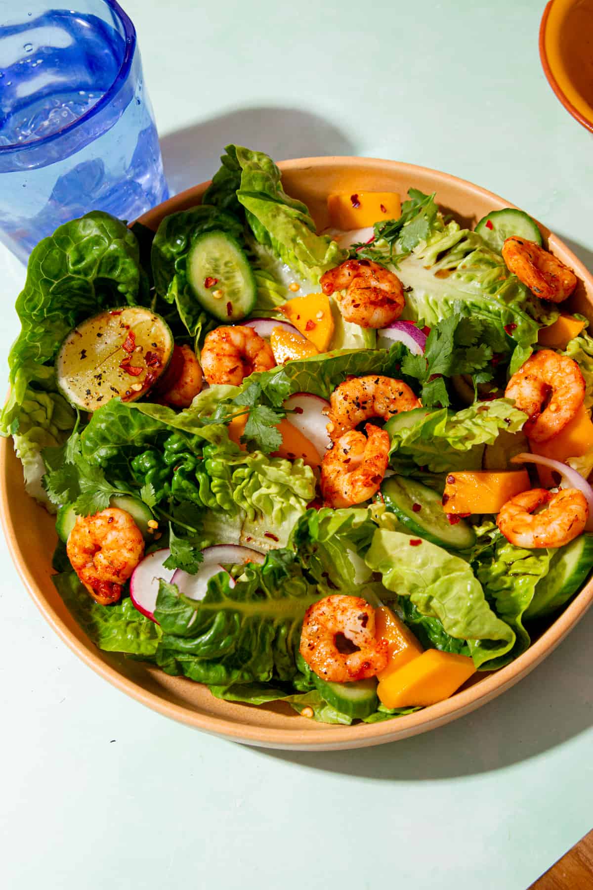 Golden browned king prawns in a bowl of salad with sliced cucumber, radished and red onion next to a blue glass of water.