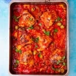 Browned chicken thighs in a tomato sauce with green olives and herbs in a baking tray on a blue background.