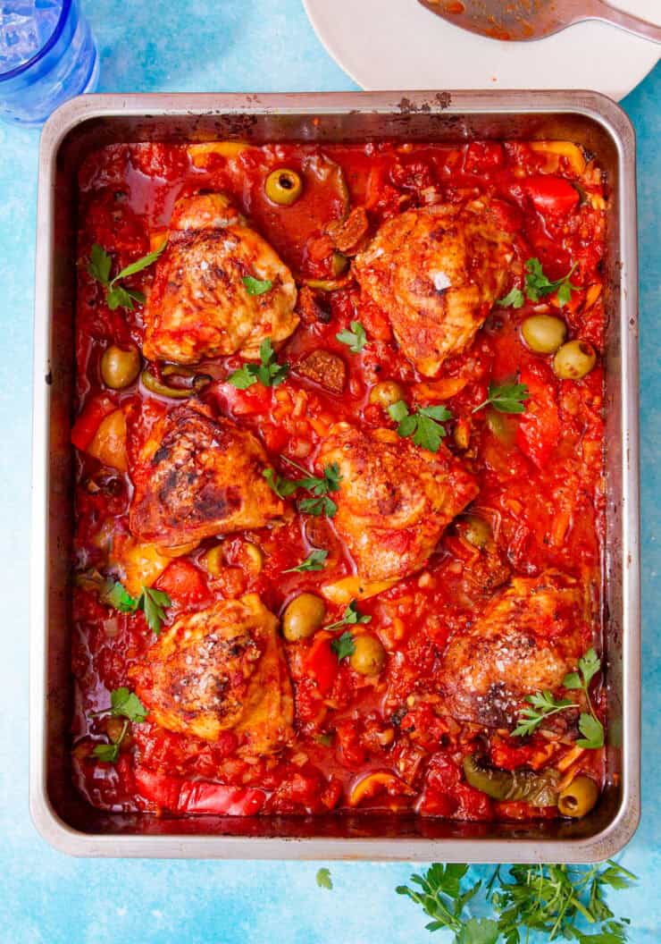 Browned chicken thighs in a tomato sauce with green olives and herbs in a baking tray on a blue background.