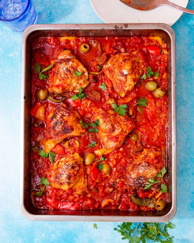 Browned chicken thighs in a tomato sauce with green olives and herbs in a baking tray on a blue background.