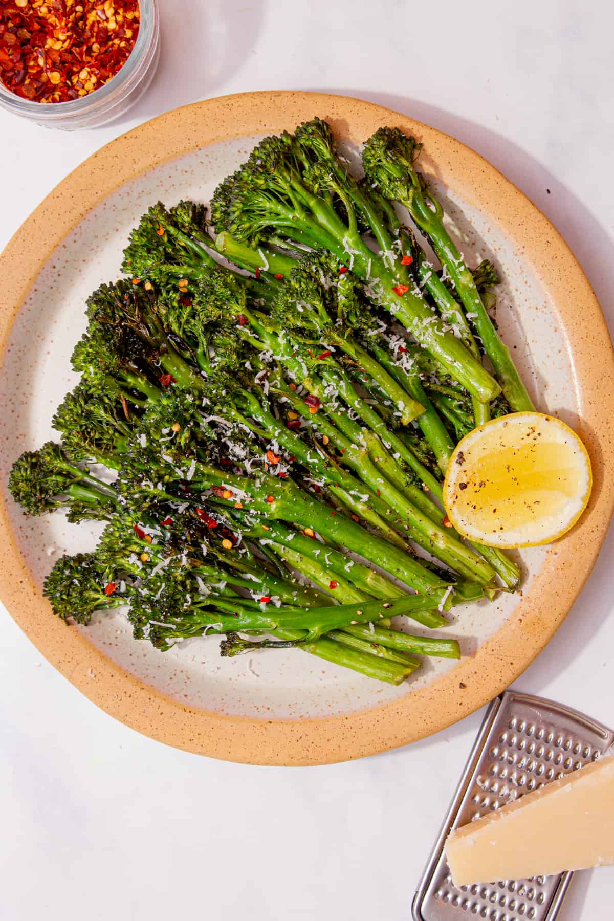Tenderstem broccoli displayed on a plate with a wedge of lemon and a small bowl of chilli flakes in partial view.