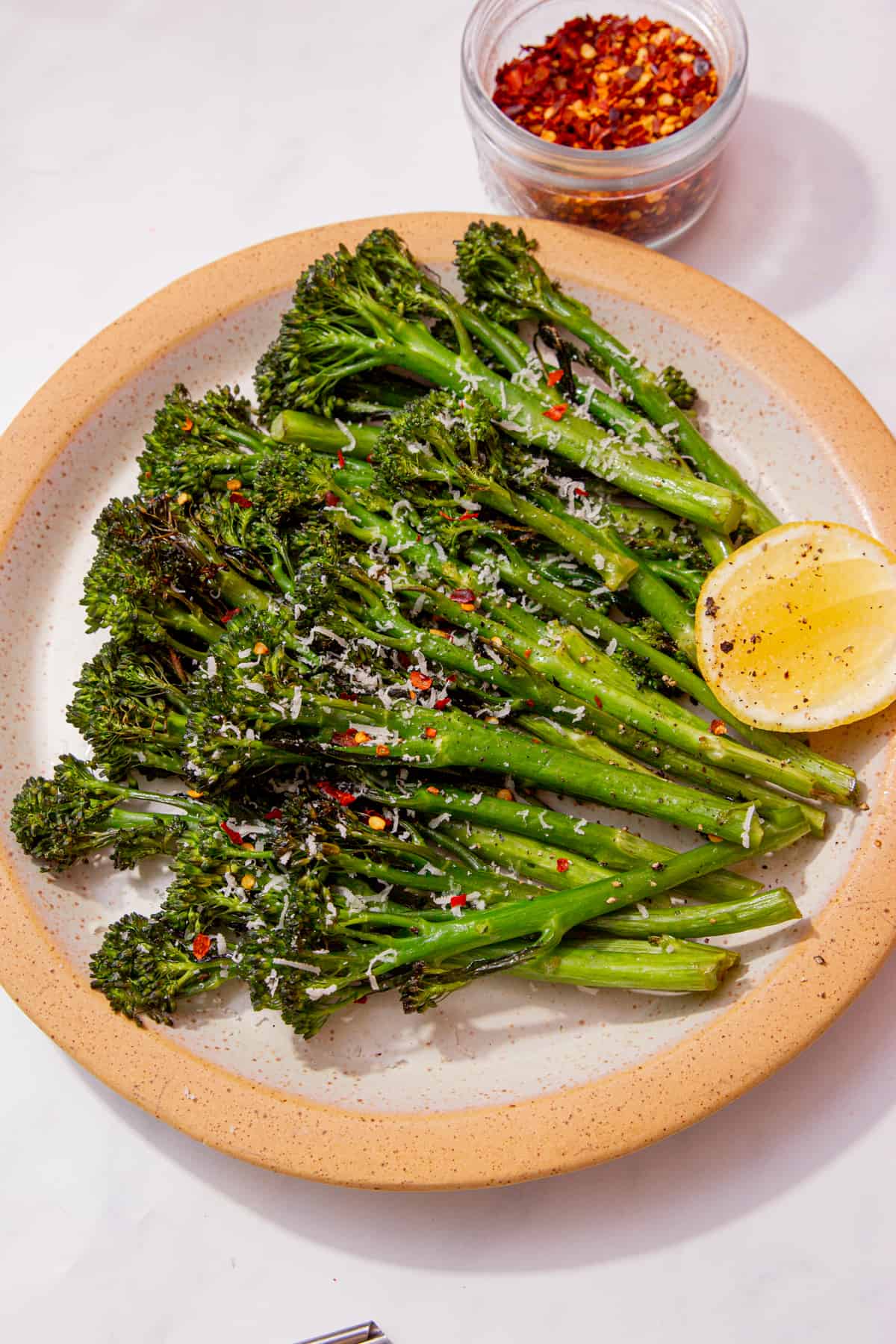 Tenderstem broccoli displayed on a plate with a wedge of lemon and a small bowl of chilli flakes behind the plate.