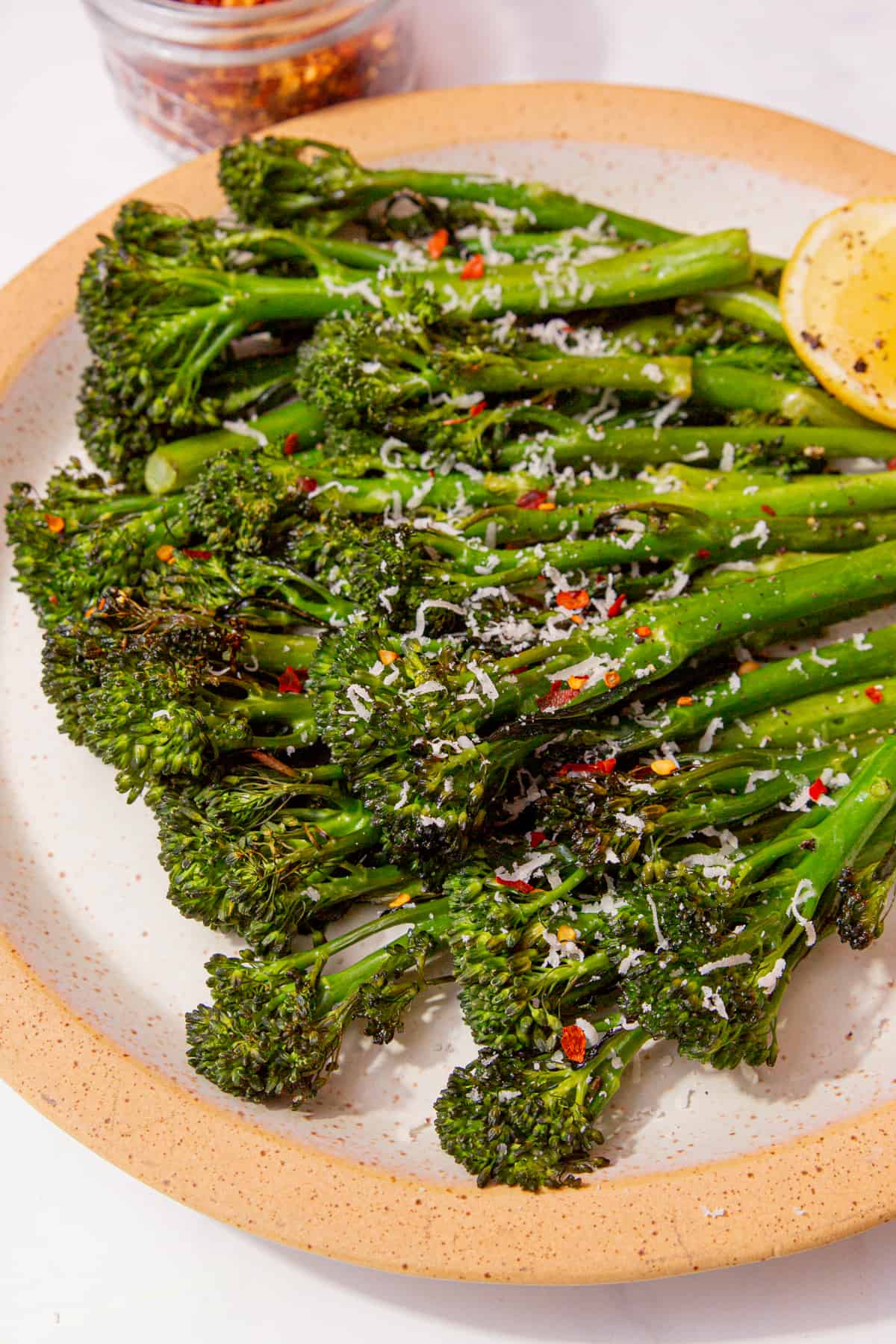 Tenderstem broccoli displayed on a plate with a wedge of lemon and topped with lots of chilli flakes and a small bowl of chilli flakes in partial view.