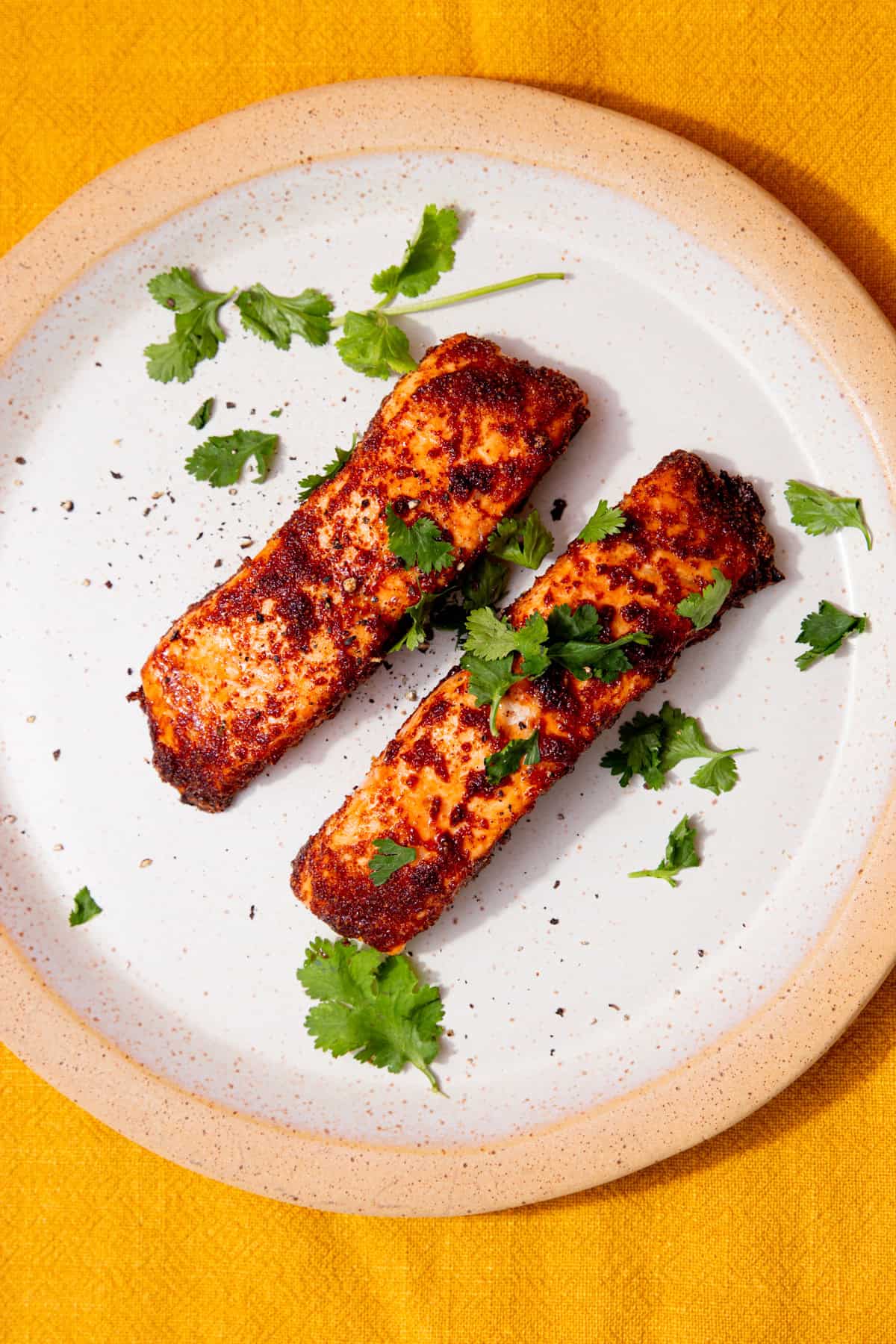 2 cooked salmon fillets with a coating of darker browned seasoning on a plate and some fresh coriander on a mustrad background.