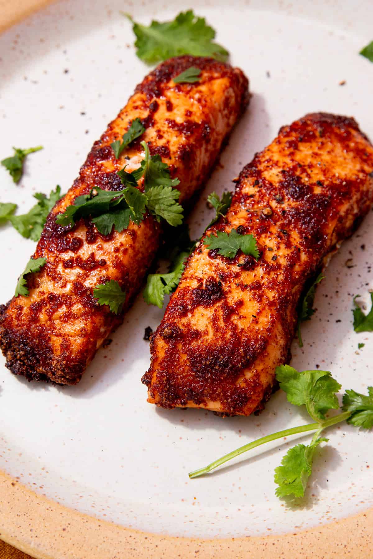 2 cooked salmon fillets with a coating of darker browned seasoning on a plate and some fresh coriander.
