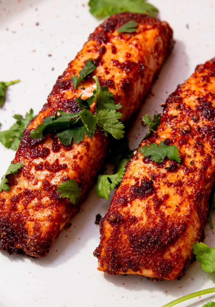 2 cooked salmon fillets with a coating of darker browned seasoning on a plate topped with some fresh coriander.