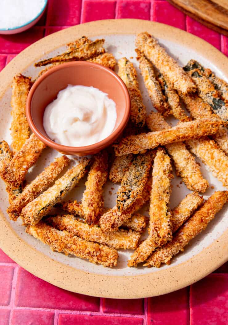 Golden browned zuccini fries on a plate with a little bowl of mayonnaise.