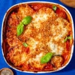 A white square dish with aubergine parmigiana topped with a golden browned cheesy topping and browned breadcrumbs next to a wooden board with grated cheese and basil leaves next to a little bowl of peppercorns.