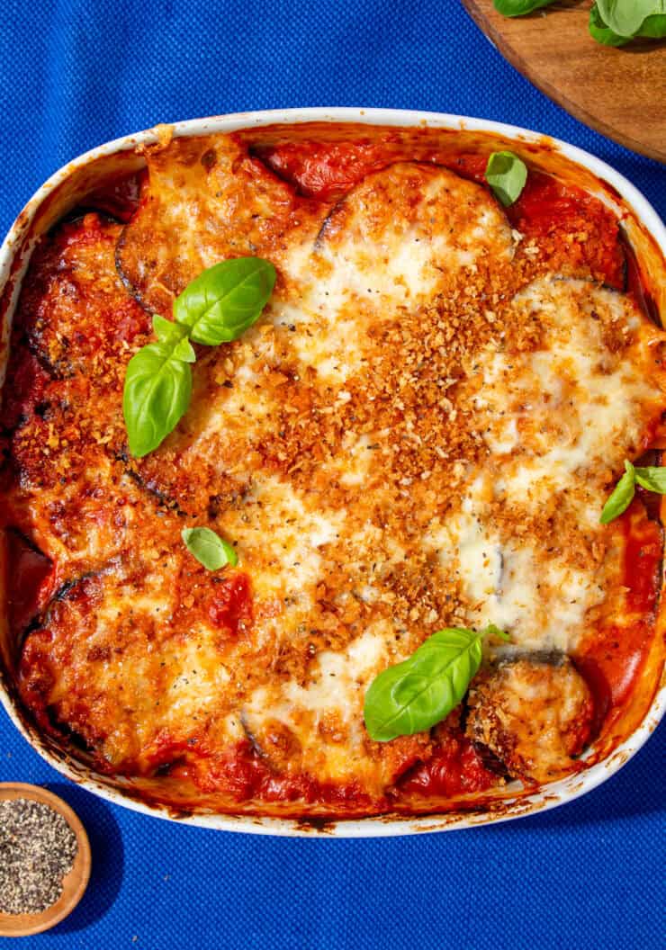 A white square dish with aubergine parmigiana topped with a golden browned cheesy topping and browned breadcrumbs next to a wooden board with grated cheese and basil leaves next to a little bowl of peppercorns.