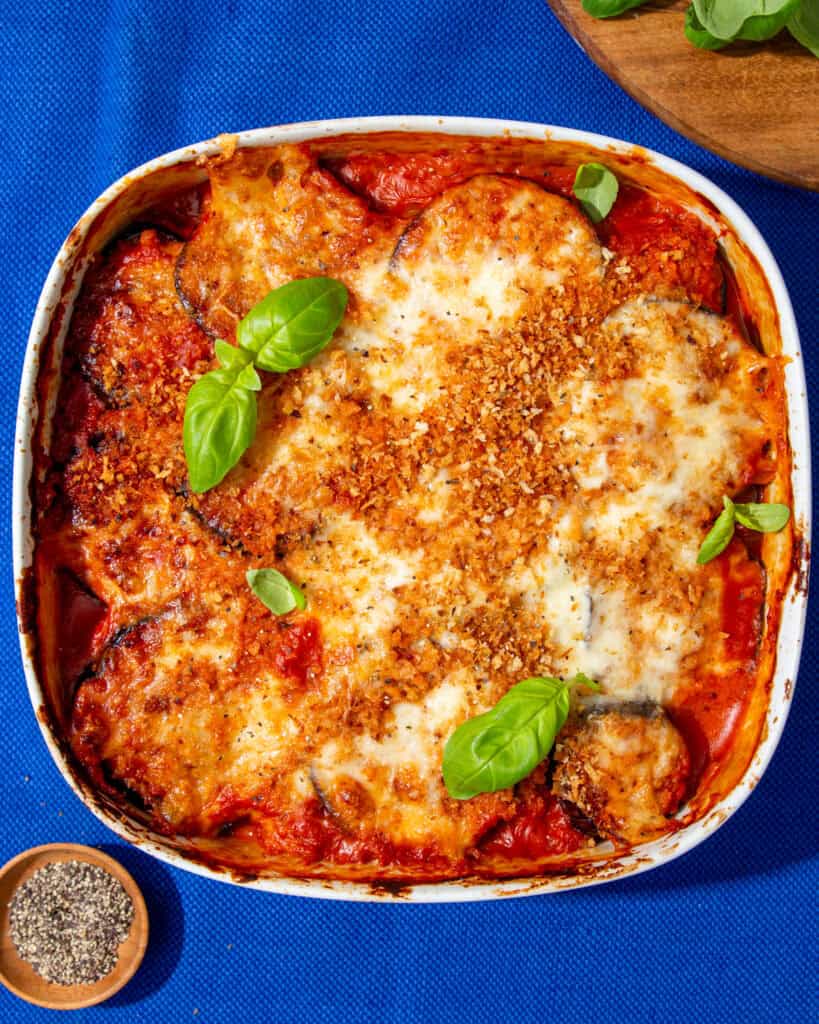 A white square dish with aubergine parmigiana topped with a golden browned cheesy topping and browned breadcrumbs next to a wooden board with grated cheese and basil leaves next to a little bowl of peppercorns.