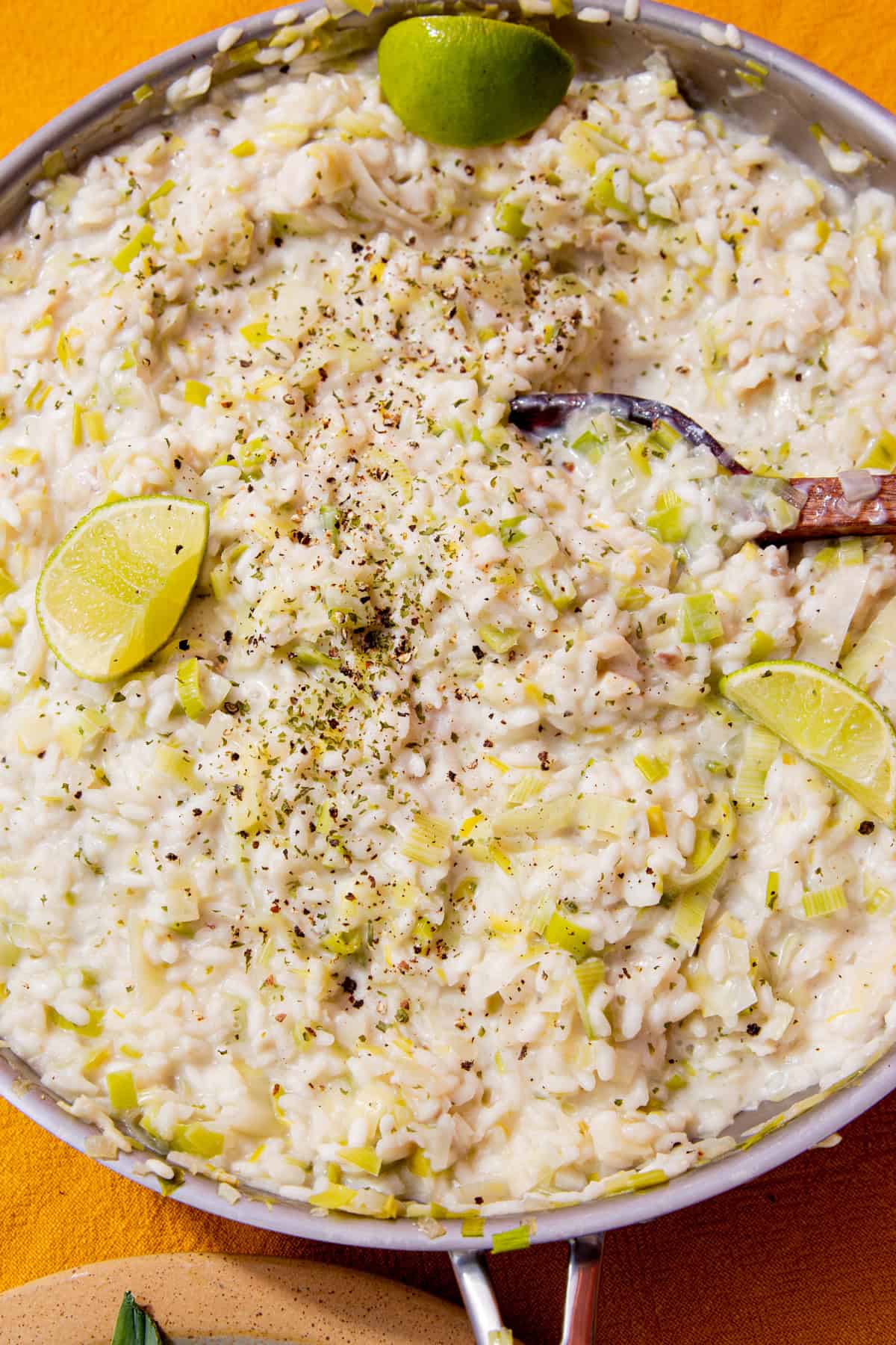 Fish risotto with leeks topped with lime wedges and some seasoning and stirred with a wooden spoon in the pan.