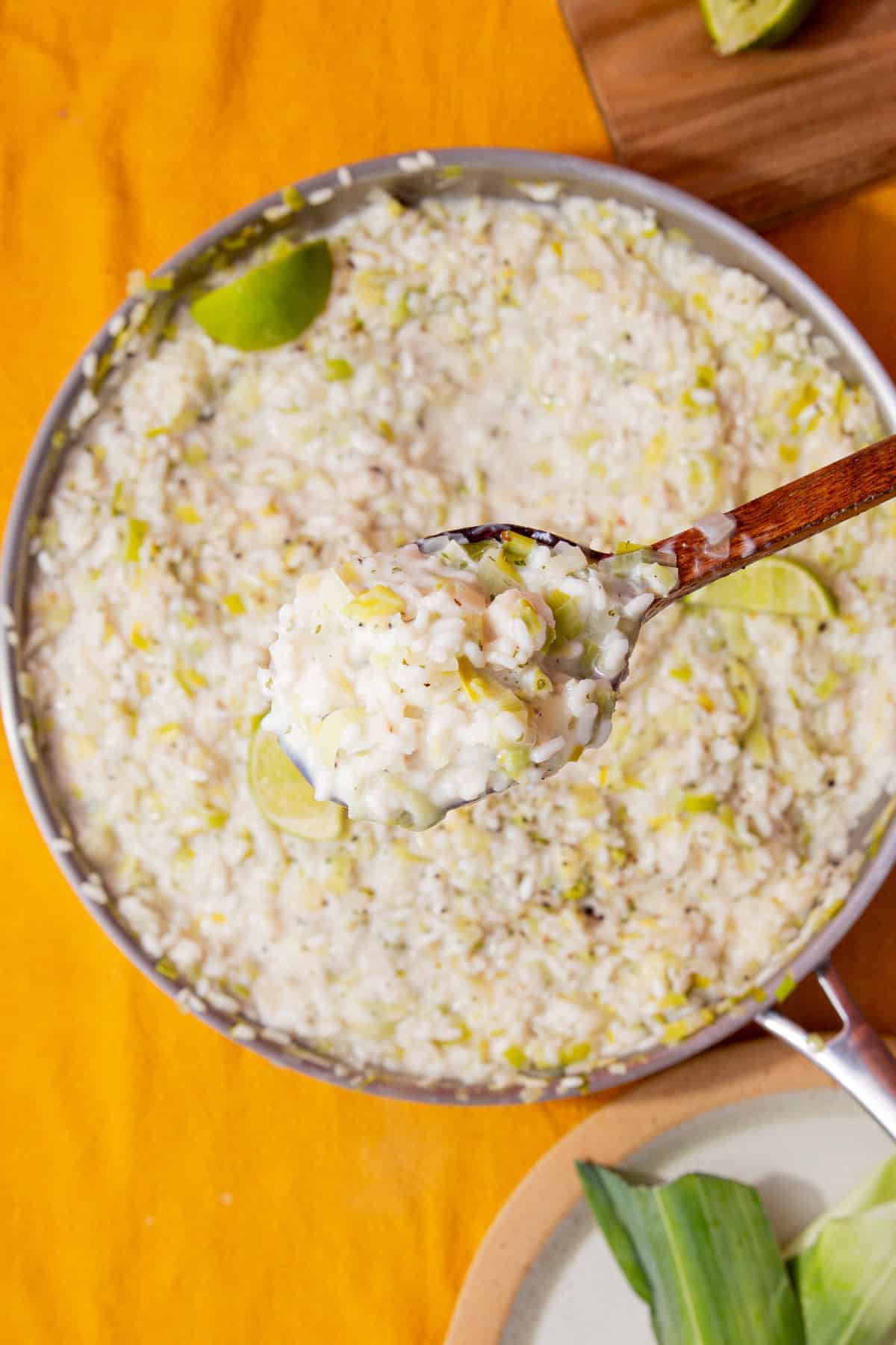 Fish risotto with leeks topped with lime wedges and some seasoning with a wooden spoon with a portion lifted up closer to view.