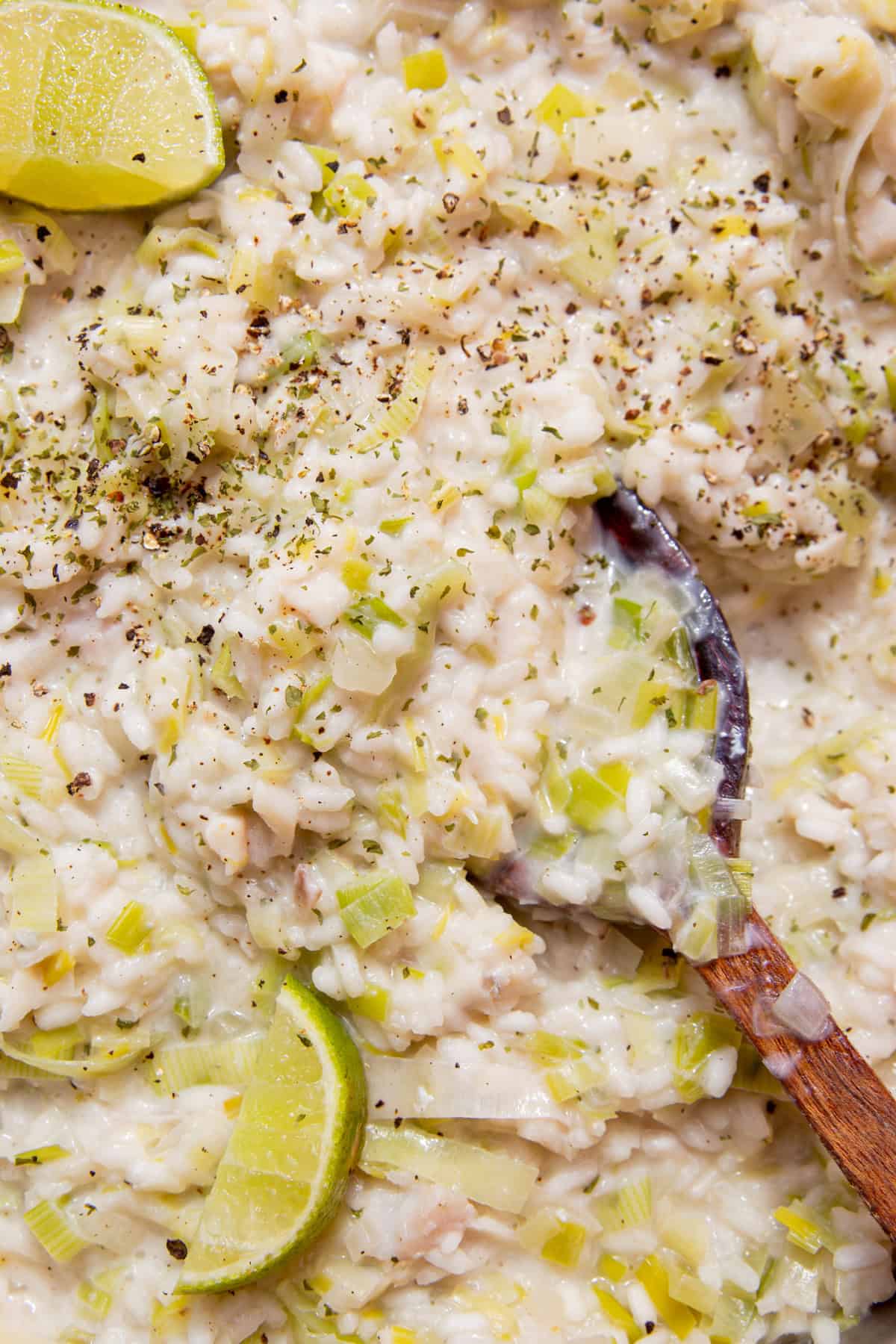 Fish risotto with leeks topped with lime wedges and some seasoning and stirred with a wooden spoon showing the moist rice mixture.