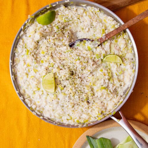 A large stainless teel pan with fish risotto topped with lime wedges, stirred with a wooden spoon on a mustrad background with a plate with leeks in partial view.