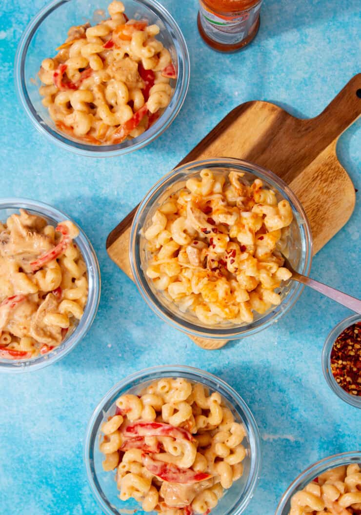 Glass meal prep containers with mac and cheese on a blue background with one dish on a wooden board.