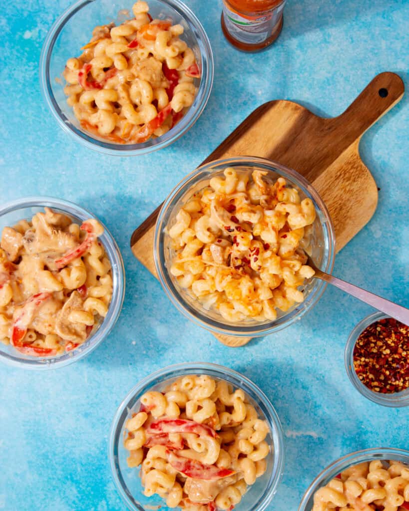 Glass meal prep containers with mac and cheese on a blue background with one dish on a wooden board.