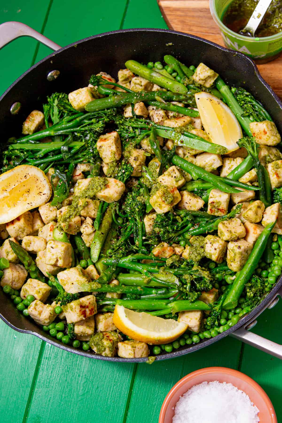 A large pan with Quorn pieces, broccoli, asparagus and peas with some lemon wedges and a green sauce.