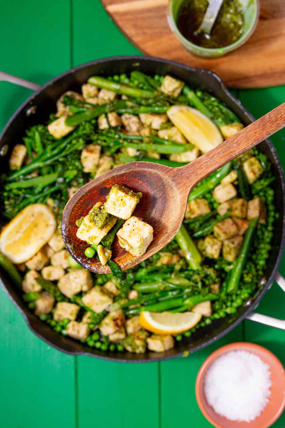 A large pan with a few Quorn pieces on a wooden spoon held up over the pan with broccoli, asparagus and peas with some lemon wedges and a green sauce next to a small bowl of salt.