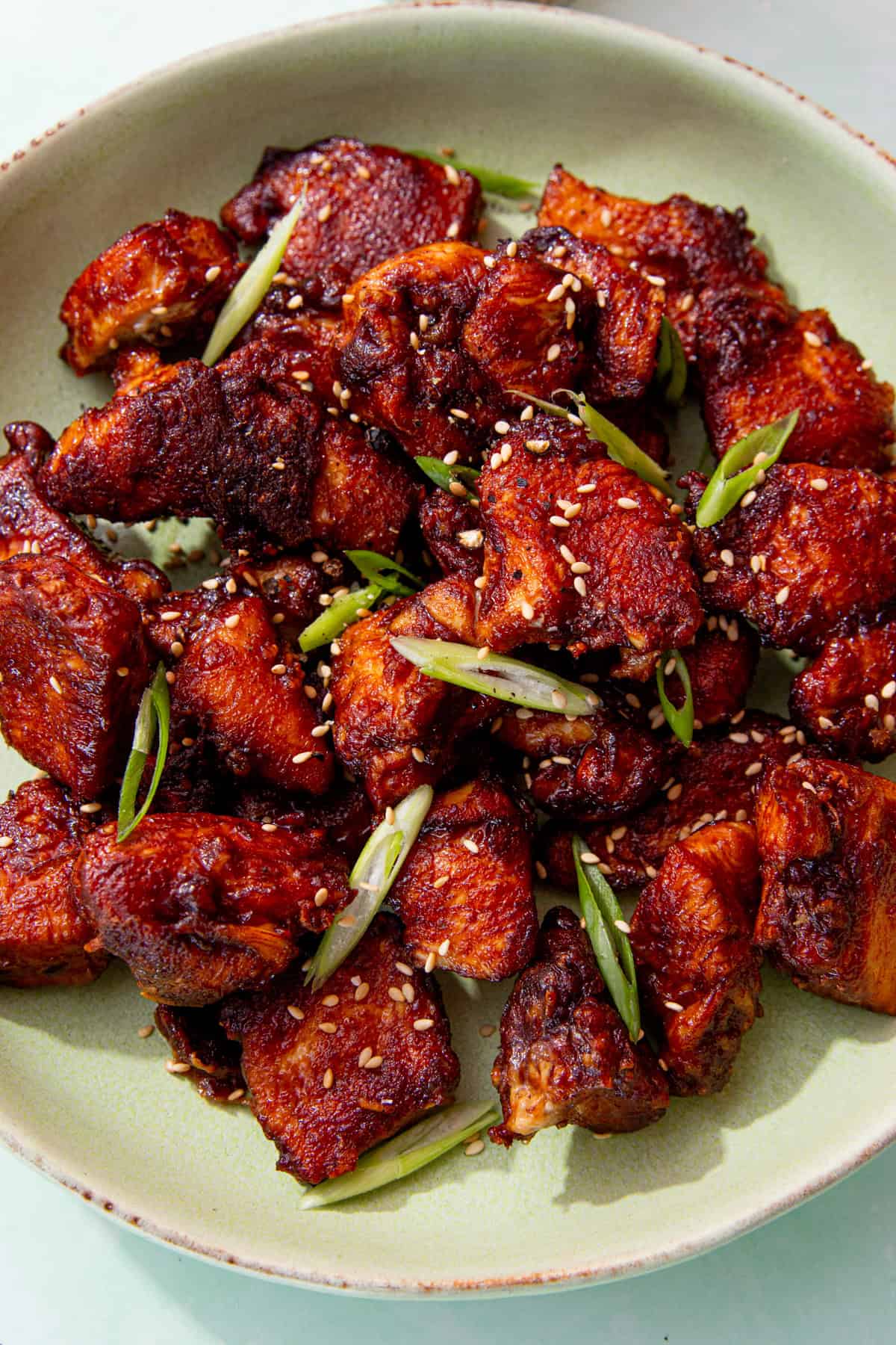 A bowl with browned soy chicken pieces topped with slices of spring onion and sesame seeds on a pale green plate.