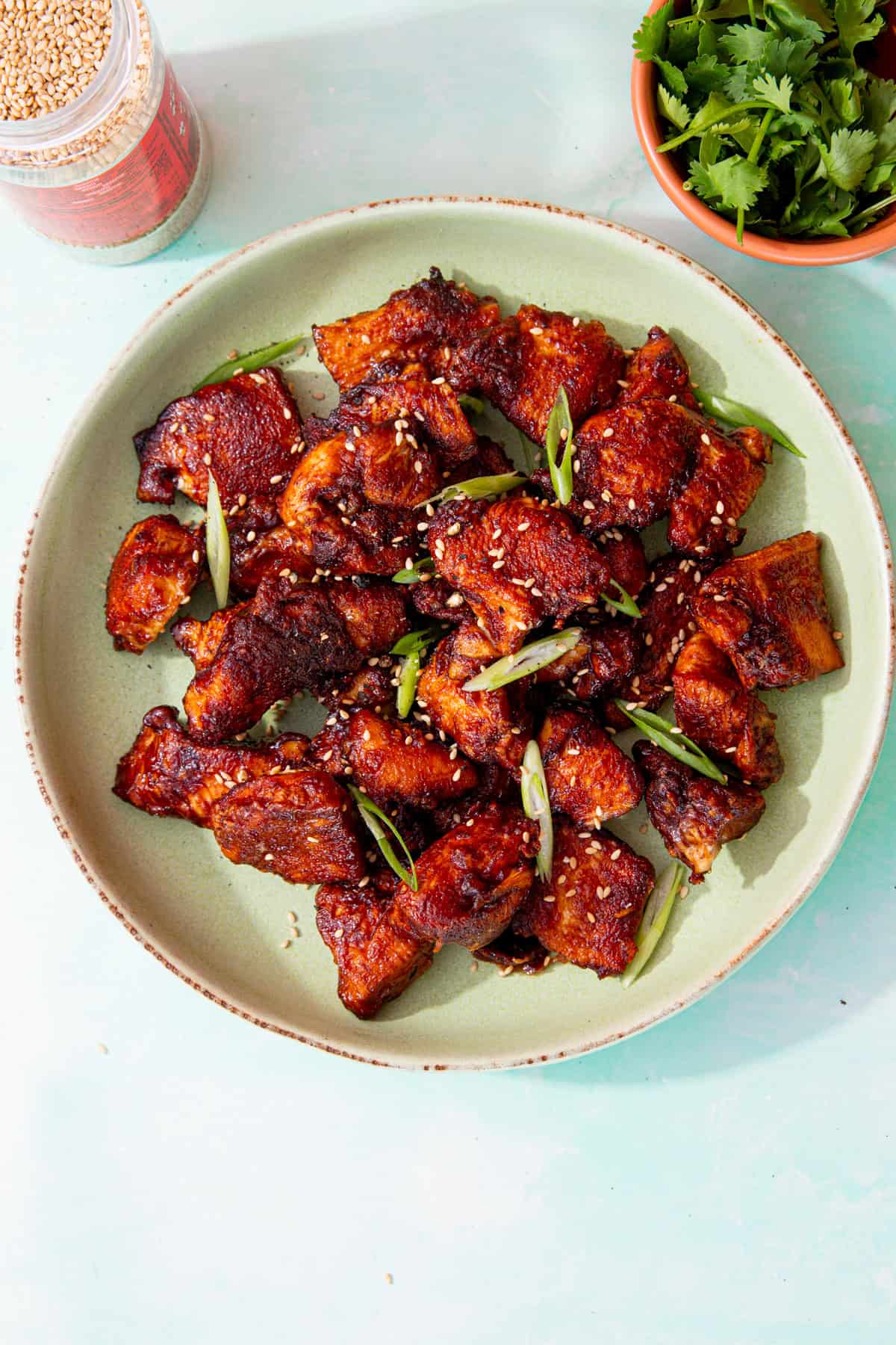 A bowl with browned soy chicken pieces topped with slices of spring onion and sesame seeds on a pale green plate with coriander and sesame seed in partial view.