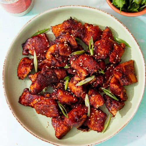 A bowl with browned soy chicken pieces topped with slices of spring onion and sesame seeds on a pale green plate next to a bowl of coriander and a container of sesame seeds.