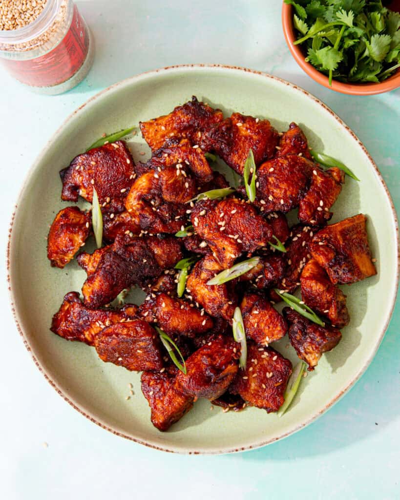 A bowl with browned soy chicken pieces topped with slices of spring onion and sesame seeds on a pale green plate next to a bowl of coriander and a container of sesame seeds.