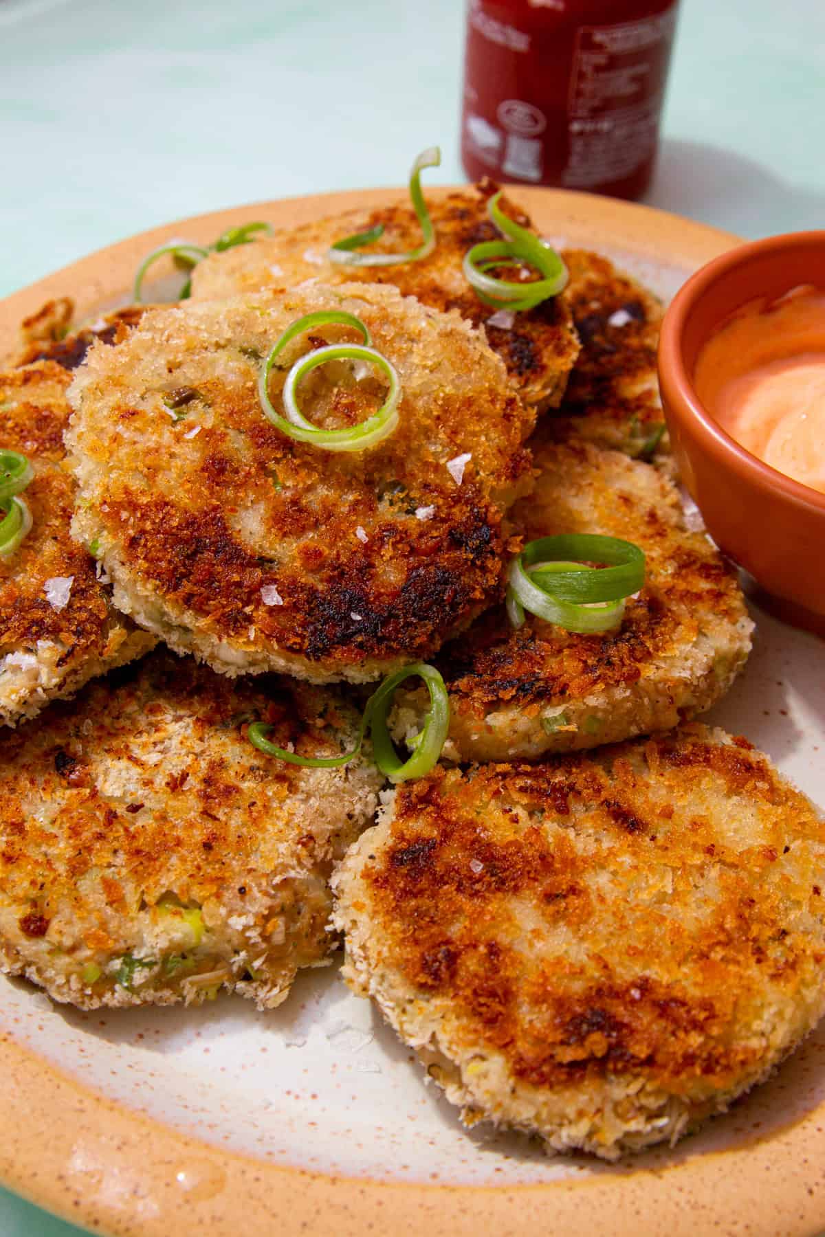 Golden browned fishcakes, piled on a plate with spirals of spring onion and a small bowl of dip and a container of sauce in partial view.