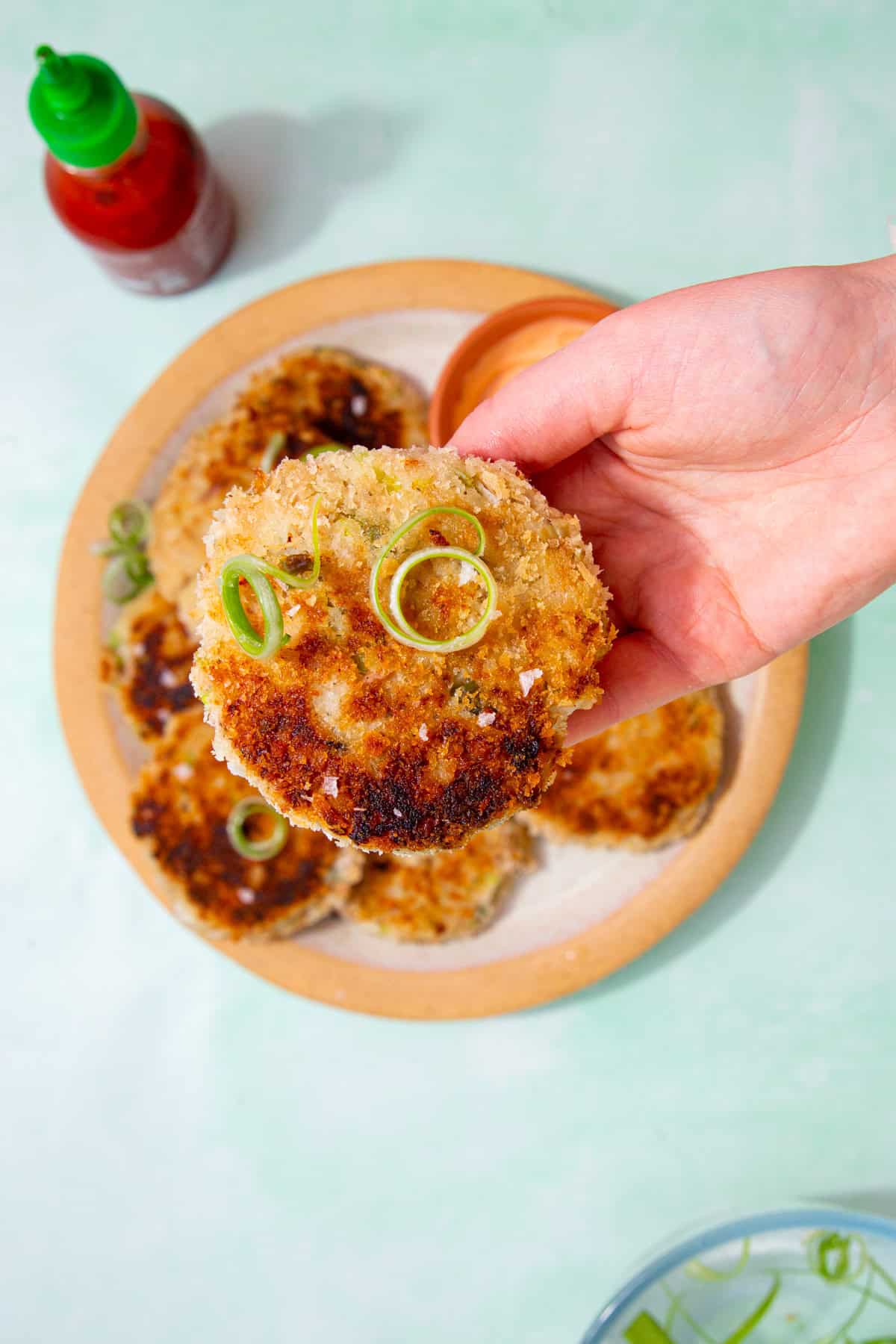 A golden browned fishcake held in a hand over a plate of fishcakes with spirals of spring onion and a sauce container next to the plate.