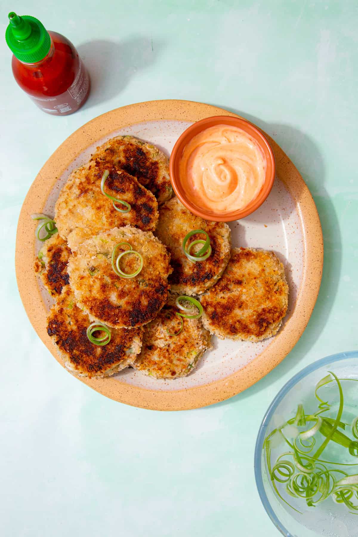 Golden browned fishcakes, piled on a plate with spirals of spring onion and a small bowl of dip and a container of sauce and a bowl with water and spirals of spring onions.