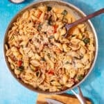 A large pan with a creamy pasta with vegetables and mince being mixed with a wooden spoon on a blue background.