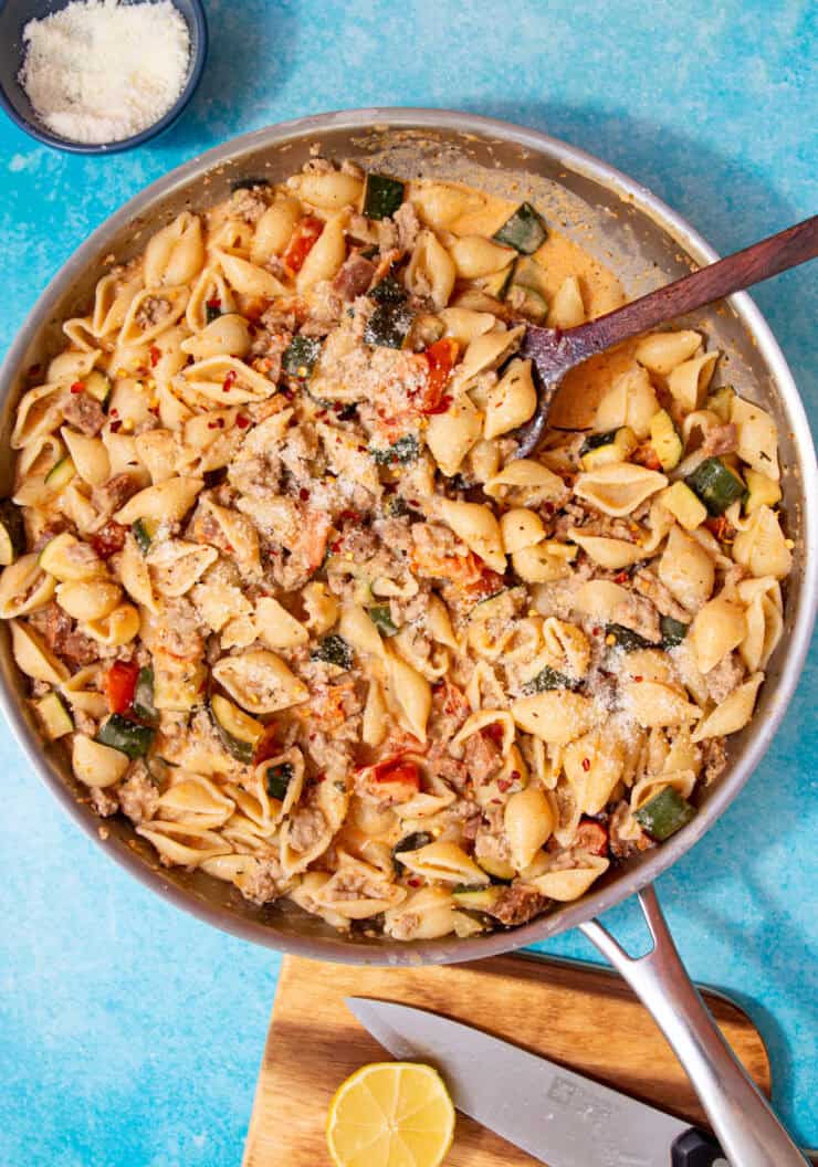 A large pan with a creamy pasta with vegetables and mince being mixed with a wooden spoon on a blue background.