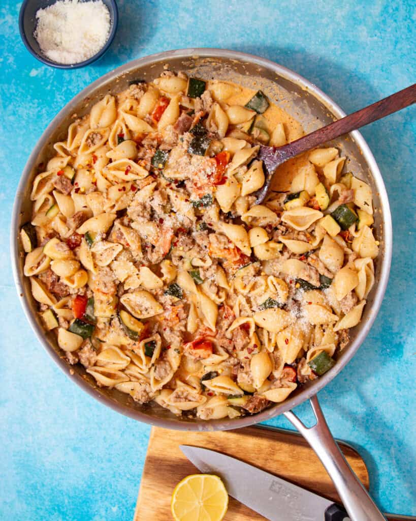 A large pan with a creamy pasta with vegetables and mince being mixed with a wooden spoon on a blue background.