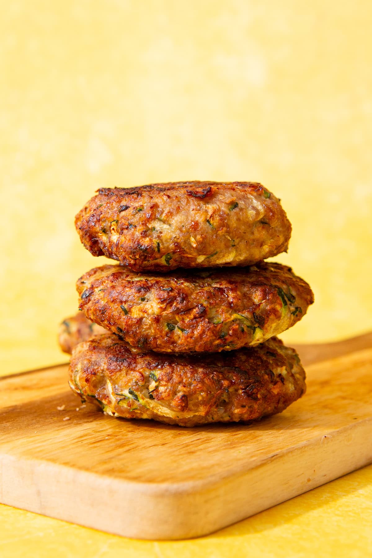 3 burgers piled on top of each other on a wooden board with a pale yellow background.