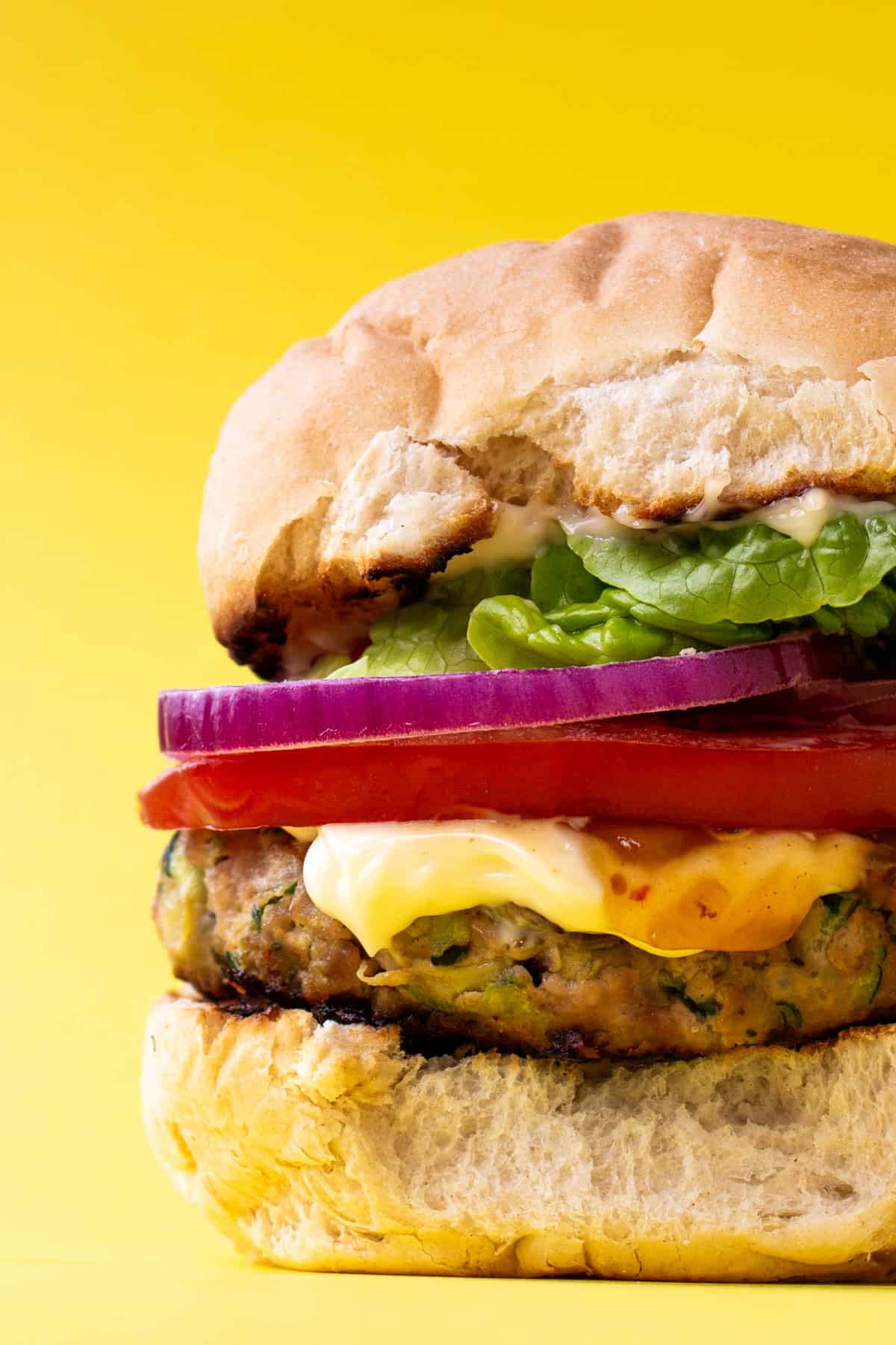 A burger in a bun with lettuce, mayonnaise, red onion and sliced tomato on a yellow background.