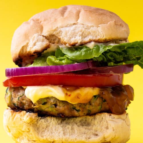 A burger in a bun with lots of filling including lettuce, tomato, mayonnaise and red onion on a yellow background.