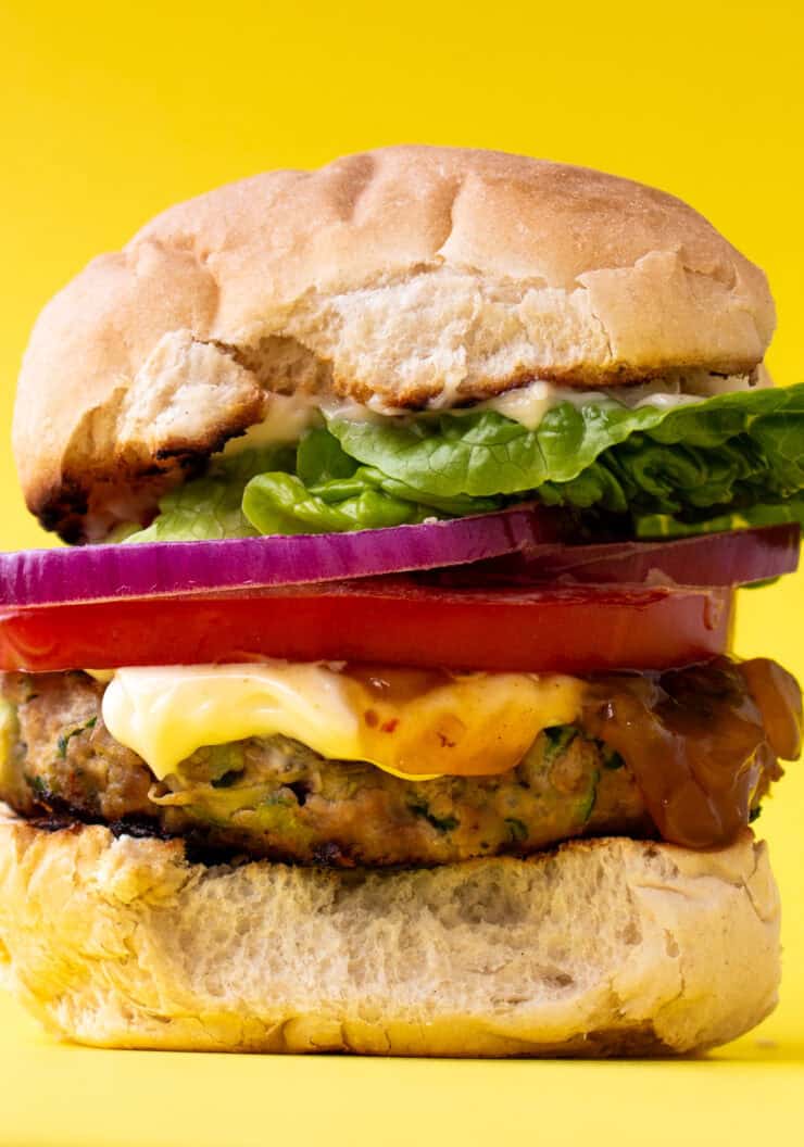 A burger in a bun with lots of filling including lettuce, tomato, mayonnaise and red onion on a yellow background.