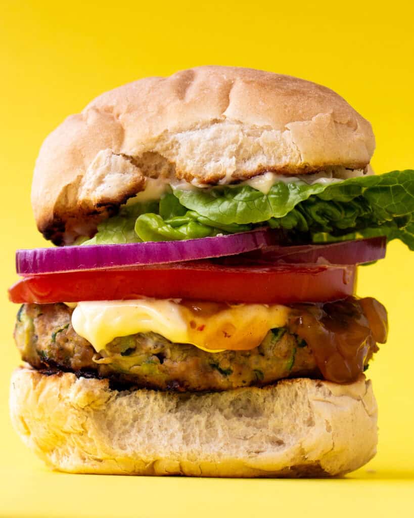 A burger in a bun with lots of filling including lettuce, tomato, mayonnaise and red onion on a yellow background.